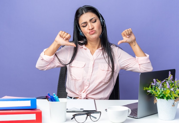 Foto giovane bella donna in abiti casual che indossa le cuffie con microfono che sembra sicura di sé mentre si siede al tavolo con il computer portatile su blue