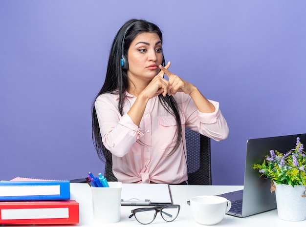 Young beautiful woman in casual clothes wearing headset with microphone showing stop gesture crossing fingers sitting at the table with laptop over blue wall working in office