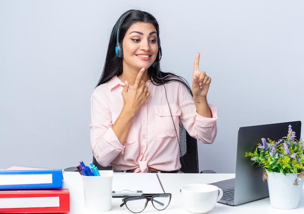 Giovane bella donna in abiti casual con cuffie e microfono seduto al tavolo con il computer portatile guardando lo schermo che mostra le dita indice sorridente sul muro bianco che lavora in ufficio