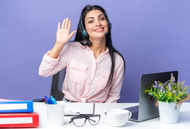 Giovane bella donna in abiti casual con cuffie e microfono sorridente fiducioso agitando con la mano seduto al tavolo con il computer portatile su sfondo blu lavorando in ufficio