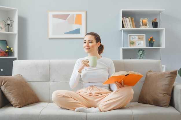 Young beautiful woman in casual clothing sitting on a couch at home interior holding a mug drinking tea and reading a book looking confident spending weekend at home