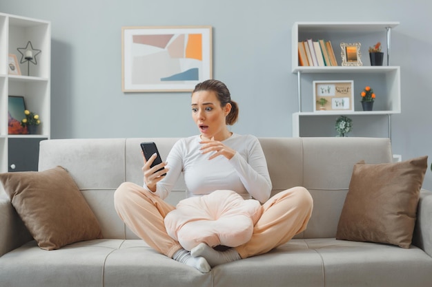 Young beautiful woman in casual clothing sitting on a couch at home interior holding smartphone looking amazed and shocked while looking at the screen of her mobile phone