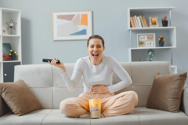 Photo young beautiful woman in casual clothing sitting on a couch at home interior with bucket of popcorn holding remote watching comedy happy and excited laughing out touching her belly spending time