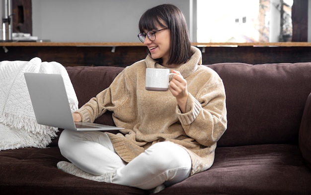 Young beautiful woman in a cozy sweater with a mug of drink works at a laptop. Remote work at home and freelancing.