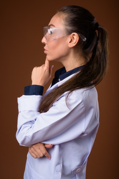 Young beautiful woman doctor wearing protective glasses against brown background