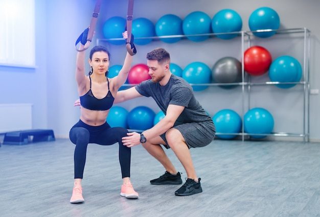 Giovane bella donna che fa esercizi con l'aiuto di una cinghia di sospensione trx con un personal trainer in palestra.