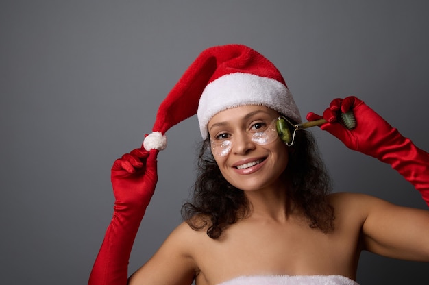 Young beautiful woman dressed as Santa uses jade roller massager for lymphatic drainage facial massage, poses on gray background with copy space for christmas advertisement of beauty salons. Skin care