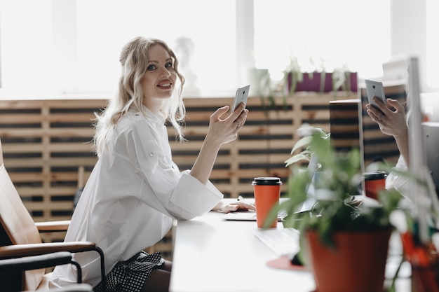 Foto la giovane bella donna vestita di camicia bianca sta lavorando con il telefono seduto alla scrivania con un computer portatile in un ufficio open space moderno e leggero