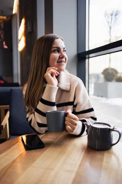 A young beautiful woman drinks coffee and looks out the window while sitting in a cafe at a table by the window. Young woman is enjoying the morning. Morning cup of invigorating coffee