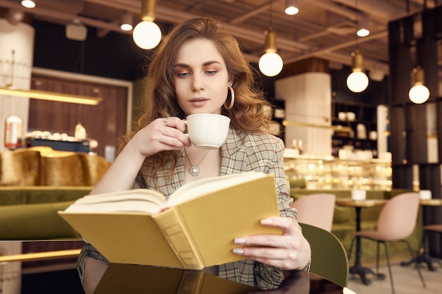 Young beautiful woman drinks coffee and reading book in cafe