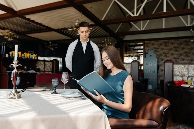 Foto una giovane e bella donna in un elegante ristorante sfoglia il menu e fa un ordine a un giovane cameriere con un elegante grembiule servizio clienti nel ristorante e negli esercizi di ristorazione