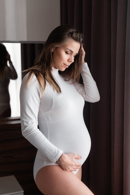 Photo young beautiful woman expecting a baby stands at a large window stroking her tummy