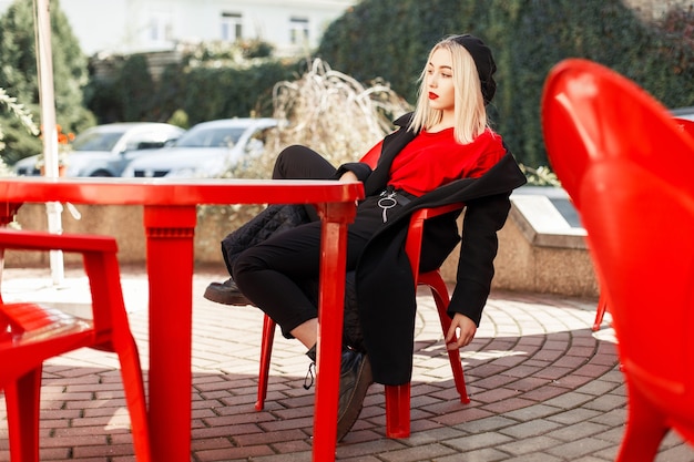 黒いコートを着たファッショナブルな秋の服を着た若い美しい女性は、カフェの赤い椅子に座っています