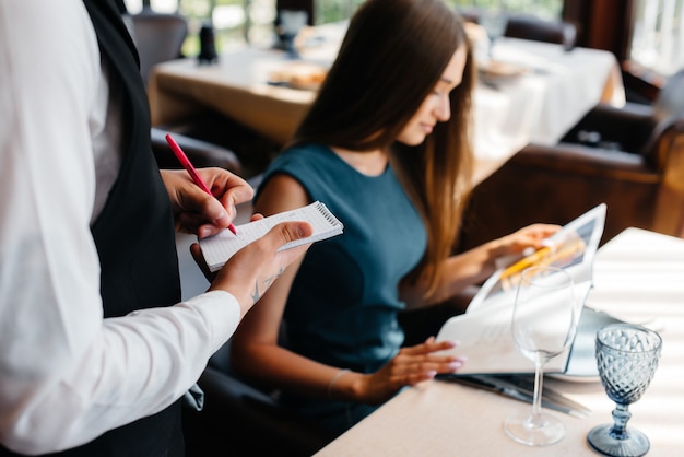 Una giovane bella donna in un raffinato ristorante guarda il menu e fa un ordine a un giovane cameriere con un grembiule alla moda. assistenza clienti.