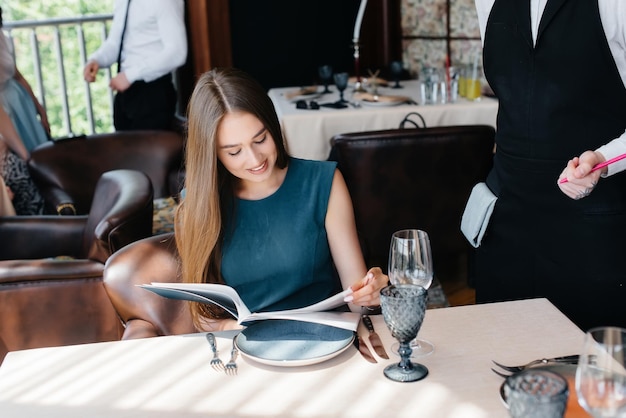 A young beautiful woman in a fine restaurant looks at the menu\
and makes an order to a young waiter in a stylish apron. customer\
service.