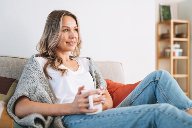 Young beautiful woman forty year with blonde long curly hair in cozy knitted grey sweater with cup of tea in hands in bright interior at the home