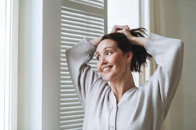 Foto giovane bella donna quarantenne con i capelli bruni in accogliente cardigan a maglia vicino alla finestra a casa