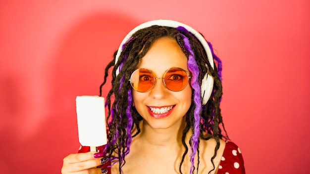 Young beautiful woman in glasses and with headphones with at ice cream red background Portrait of happy female with dreadlocks in anticipation enjoying sundae