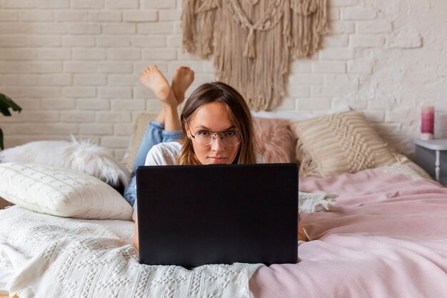 メガネの若い美しい女性は、ベッドの上のノートパソコンで動作します。
