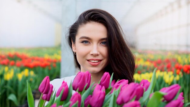 Young beautiful woman greenhouse worker holds a box with blooming tulips in her hands and smiles