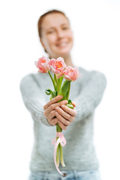 Young beautiful woman grey sweater gives pink tulips on white background. Blur portrait, selective focus. 8 March and Mother's Day.