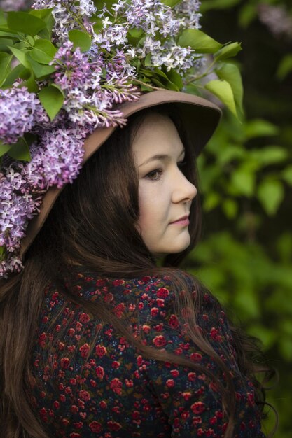 Young beautiful woman in a hat stands in front of a lilac bush Lilac wreath on the hat