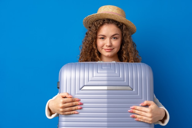 Young beautiful woman in hat on vacation with suitcase against blue background