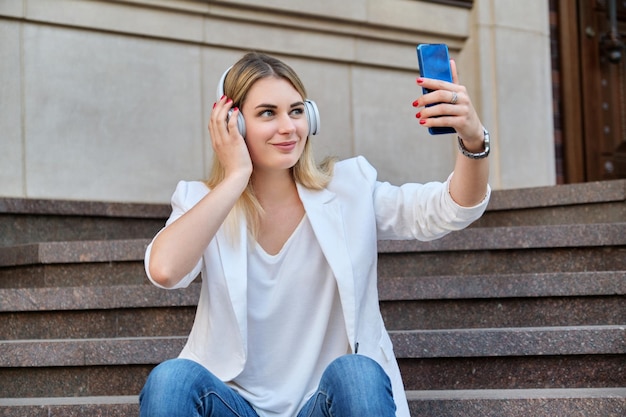 街の階段に座って休んでスマートフォンとヘッドフォンで若い美しい女性