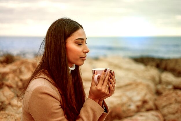 Foto giovane bellissima donna di vent'anni con lunghi capelli scuri si gode una tazza di tè alle erbe con gli occhi chiusi seduta su una roccia vicino al mare o all'oceano prendendo tempo per se stessa in mezzo alla natura per rilassarsi