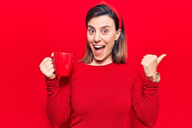 Young beautiful woman holding coffee pointing thumb up to the side smiling happy with open mouth