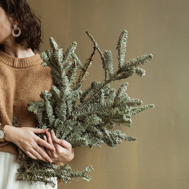 Young beautiful woman holding fir branches against olive wall