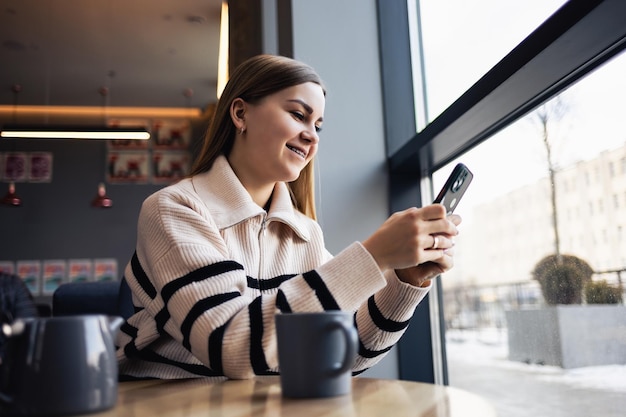 カフェテリアに座ってスマートフォンを持って見ている若い美しい女性携帯電話を使用して幸せな大学生実業家はコーヒーと笑顔を飲みます