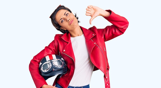 Young beautiful woman holding motorcycle helmet with angry face, negative sign showing dislike with thumbs down, rejection concept
