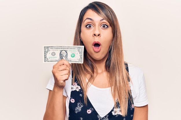 Young beautiful woman holding one dollar scared and amazed with open mouth for surprise disbelief face