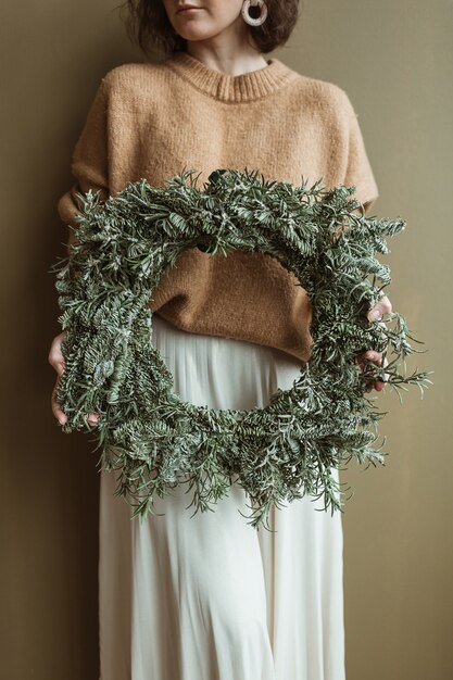 Young beautiful woman holding round wreath frame made of fir branches against olive wall