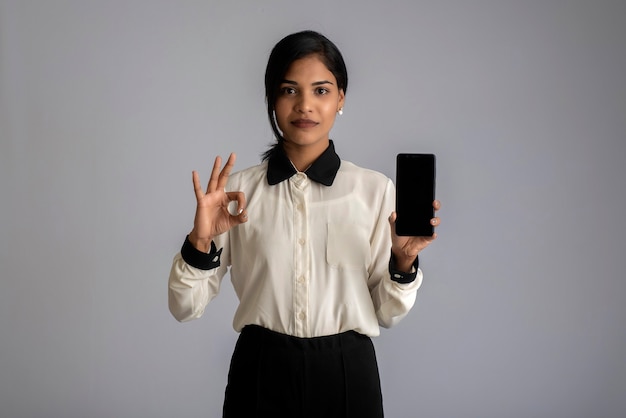 Young beautiful woman holding and showing blank screen smartphone or mobile or tablet phone on a gray wall.