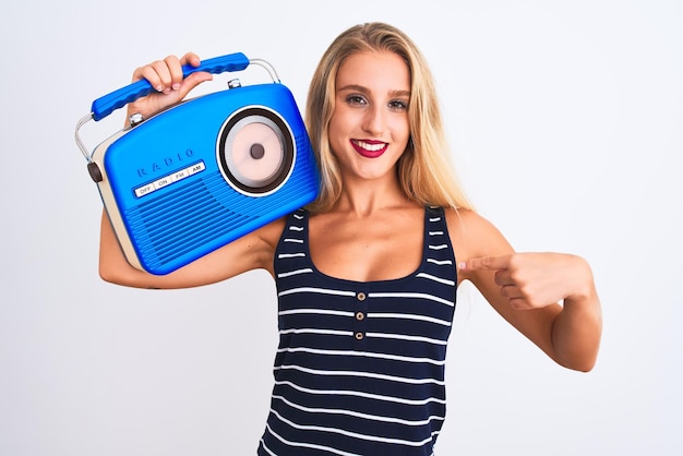 Молодая красивая женщина держит винтажное радио, стоящее на изолированном белом фоне с удивленным лицом, указывающим пальцем на себя