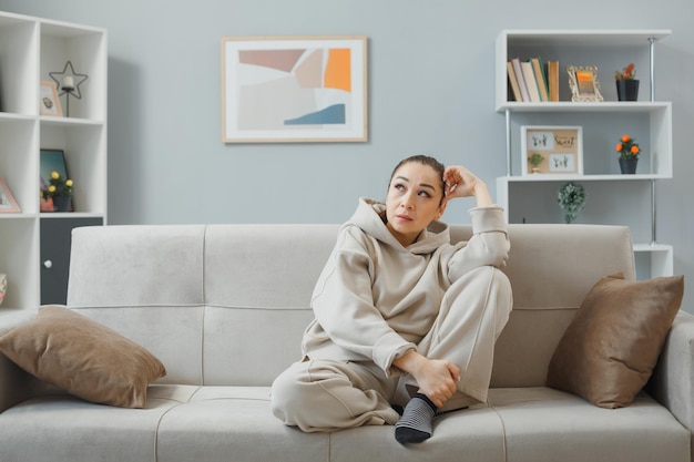 Young beautiful woman in home clothes sitting on a couch at home interior looking aside with pensive expression thinking