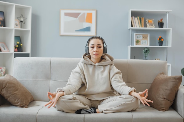 Young beautiful woman in home clothes sitting on a couch at home interior with headphones happy and positive relaxing meditating making meditation gesture with fingers