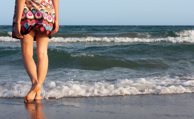 写真 夏のドレスを着た若い美しい女性は、砂浜の海の水に立って、波と潮風を楽しんでいます