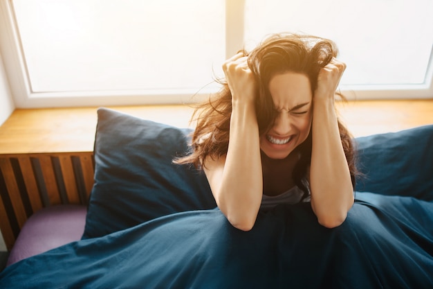 自宅の朝ベッドで若くてきれいな女性。耳に近い頭の上で手をつなぎ、痛みや頭痛に悩まされます。通りと痛みを伴う朝。