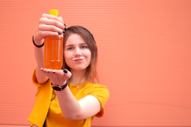 A young beautiful woman is standing on an orange wall, holding a plastic bottle of beer