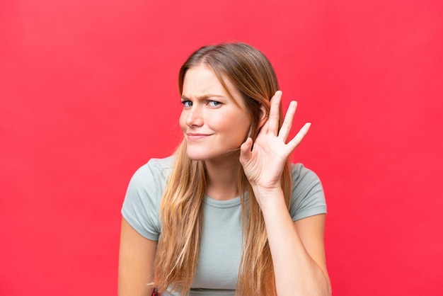 Photo young beautiful woman isolated on red background listening to something by putting hand on the ear