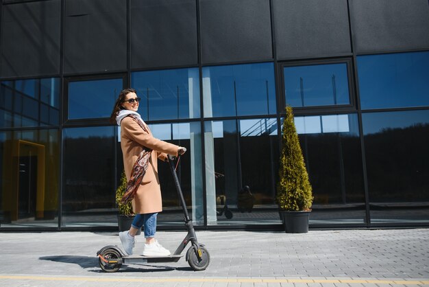 Young beautiful woman in a jacket smiles and rides an electric scooter to work 