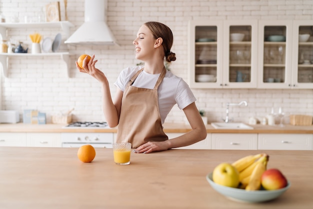 Giovane bella donna in cucina con grembiule, frutta e succo d'arancia orange