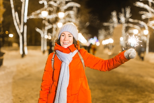 Giovane bella donna in berretto lavorato a maglia e sciarpa in piedi in città con luce bengala, sparkler. celebrazione di concetto e natale.