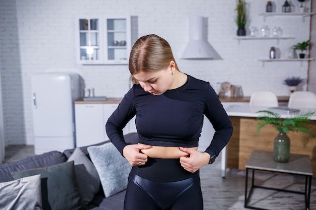 Young beautiful woman in leggings and a top shows a fold on the belly. Healthy lifestyle. The woman goes in for sports at home. Fighting excess weight at home