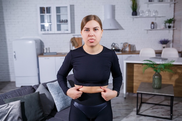 Young beautiful woman in leggings and a top shows a fold on the belly. Healthy lifestyle. The woman goes in for sports at home. Fighting excess weight at home