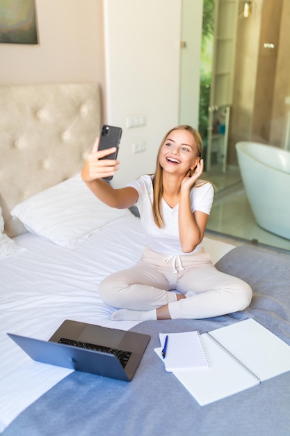 Молодая красивая женщина лежит в своей постели в постели с ноутбуком, девушка делает селфи на телефоне