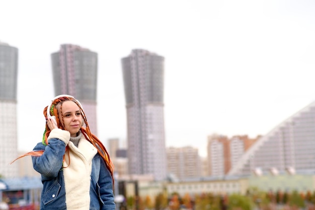 Young beautiful woman listening to music in wireless headphones on city street Vivid female with multicoloured hairstyle enjoying songs on walk in cloudy weather